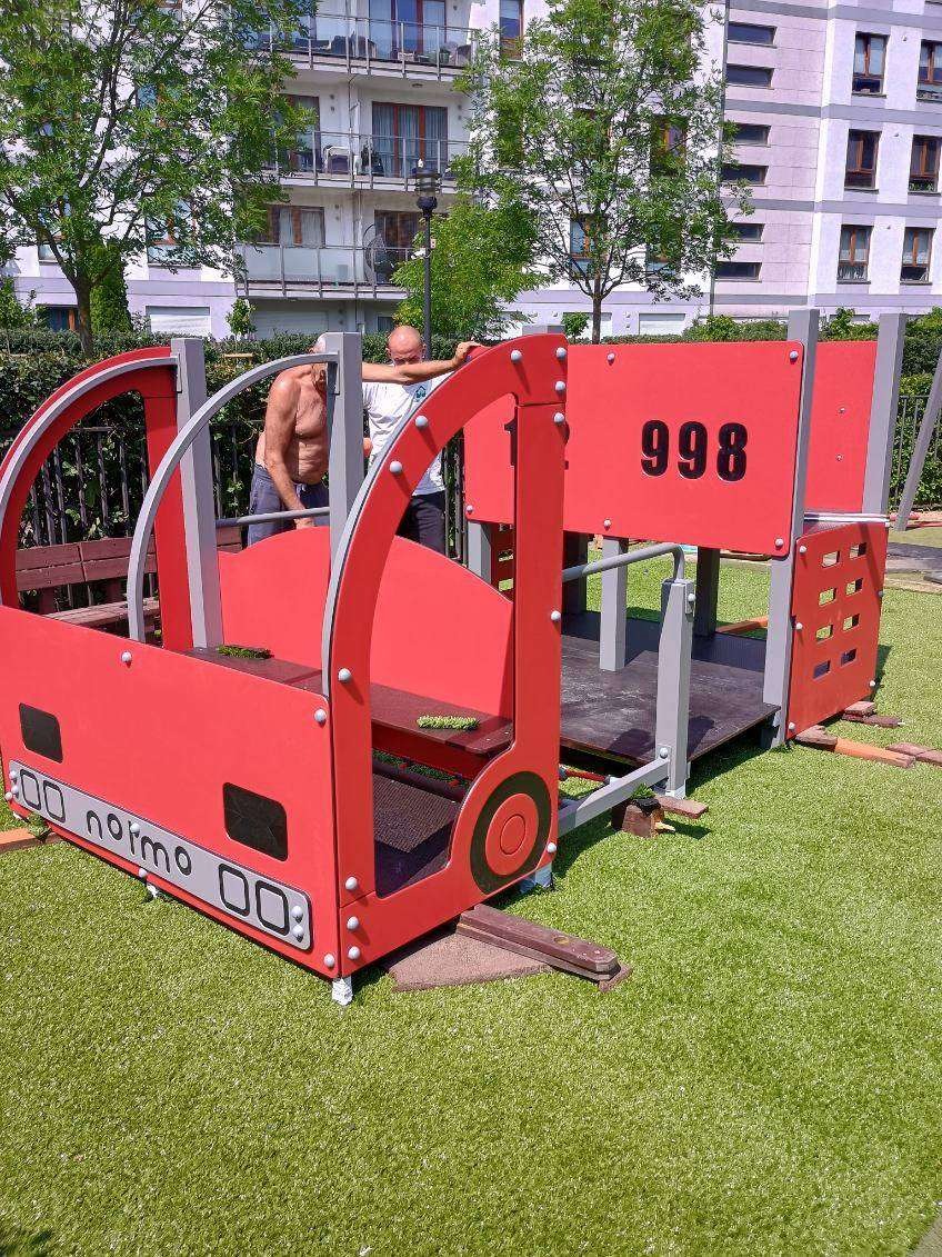 Czerwony, drewniany plac zabaw w kształcie pociągu z numerem 998 na tle budynku mieszkalnego i dwóch osób, umieszczony na sztucznej trawie.