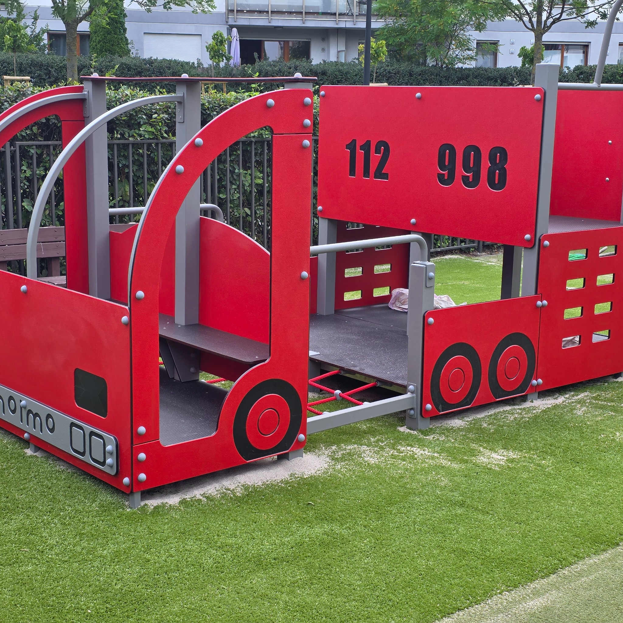 Czerwony plac zabaw w kształcie wozu strażackiego z numerami 112 i 998, na sztucznej trawie. Elementy szare, metalowe. Tło: budynek mieszkalny, krzewy.