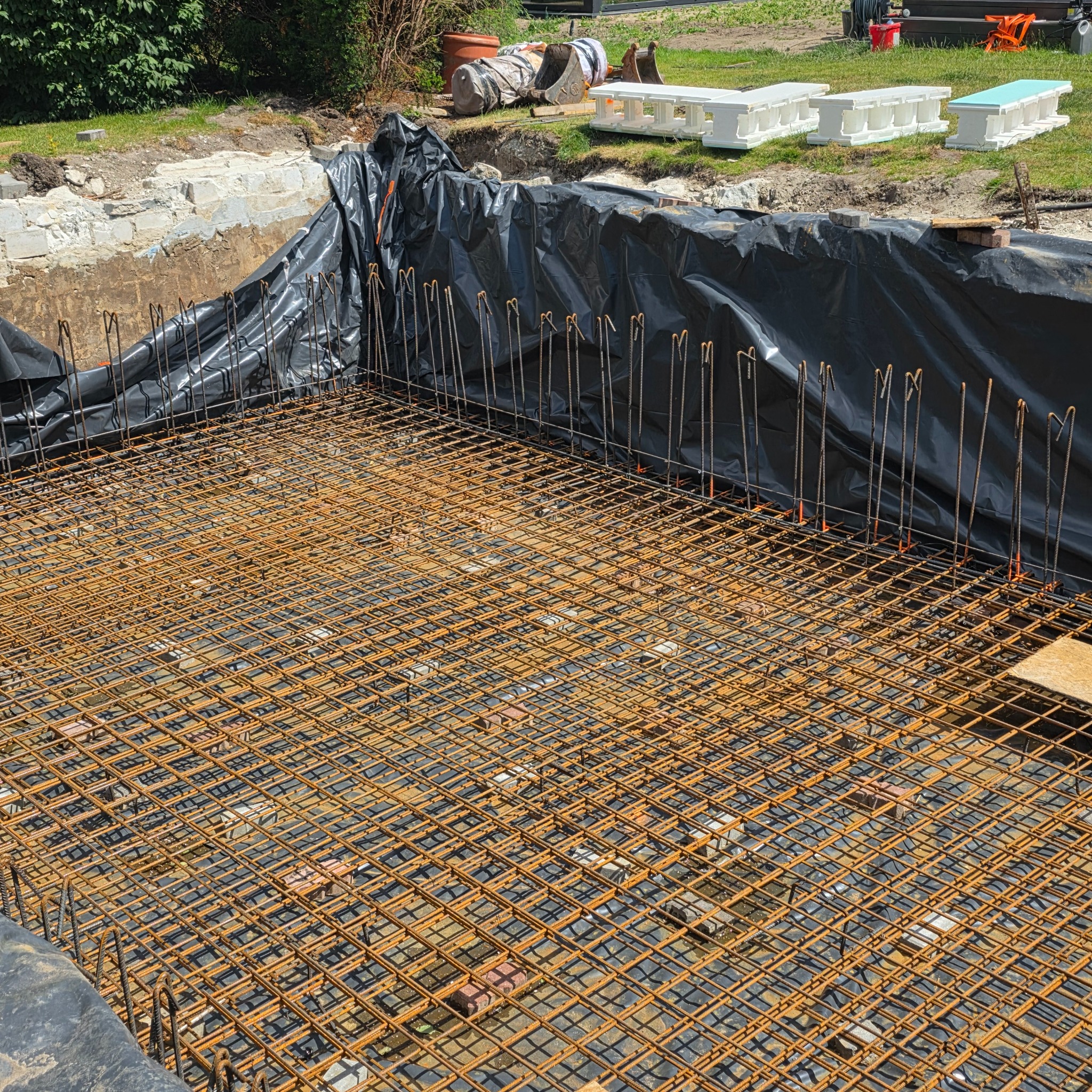 Siatka zbrojeniowa w wykopie, zabezpieczona folią, przygotowana pod wylewkę fundamentów. Widoczne narzędzia i elementy konstrukcyjne w tle.
