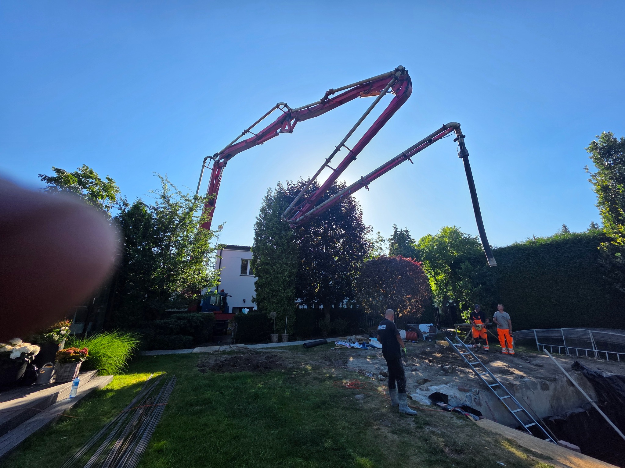 Betonomieszarka z wysięgnikiem nad wykopem fundamentowym; pracownicy w odblaskowych ubraniach, zieleń ogrodu i błękitne niebo w tle.