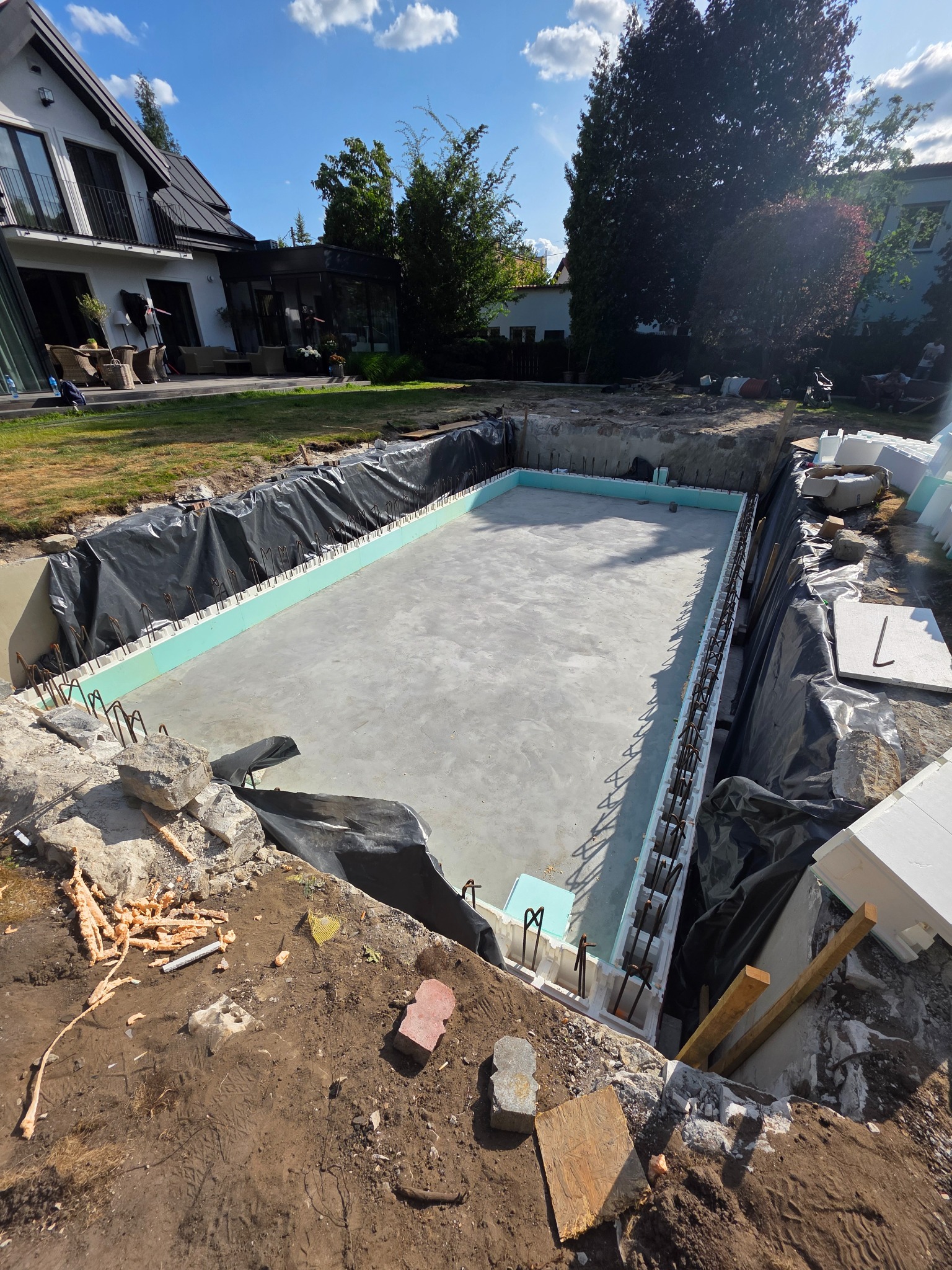Wykop pod basen w ogrodzie, betonowa płyta fundamentowa, szalunek z izolacją termiczną, folia ochronna. Widok na dom i drzewa w tle, słoneczny dzień.