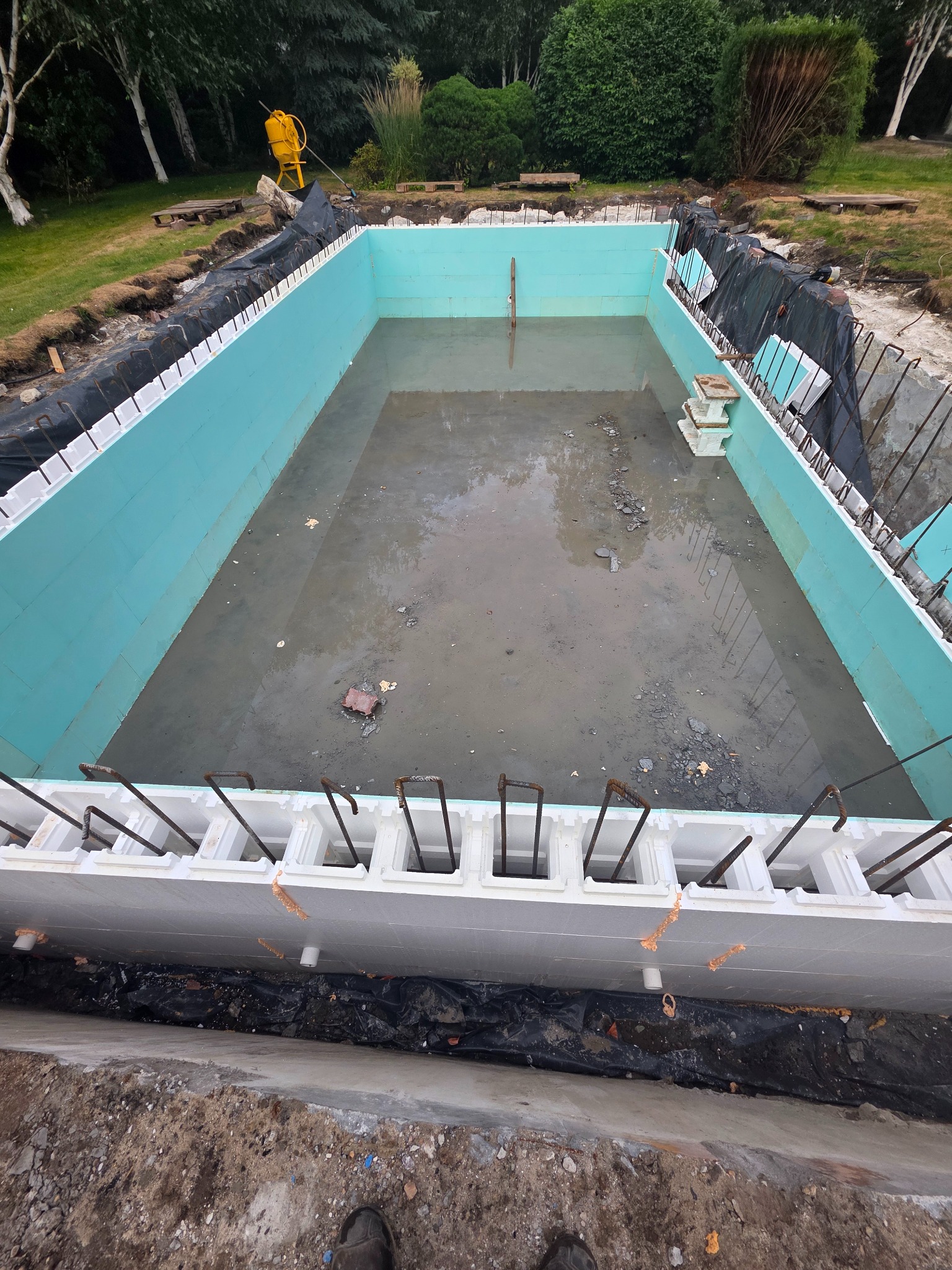 Budowa basenu w ogrodzie: widok z góry na wykop z szalunkiem z pustaków styropianowych, wypełniony wodą. W tle żółta betoniarka i zieleń ogrodu.