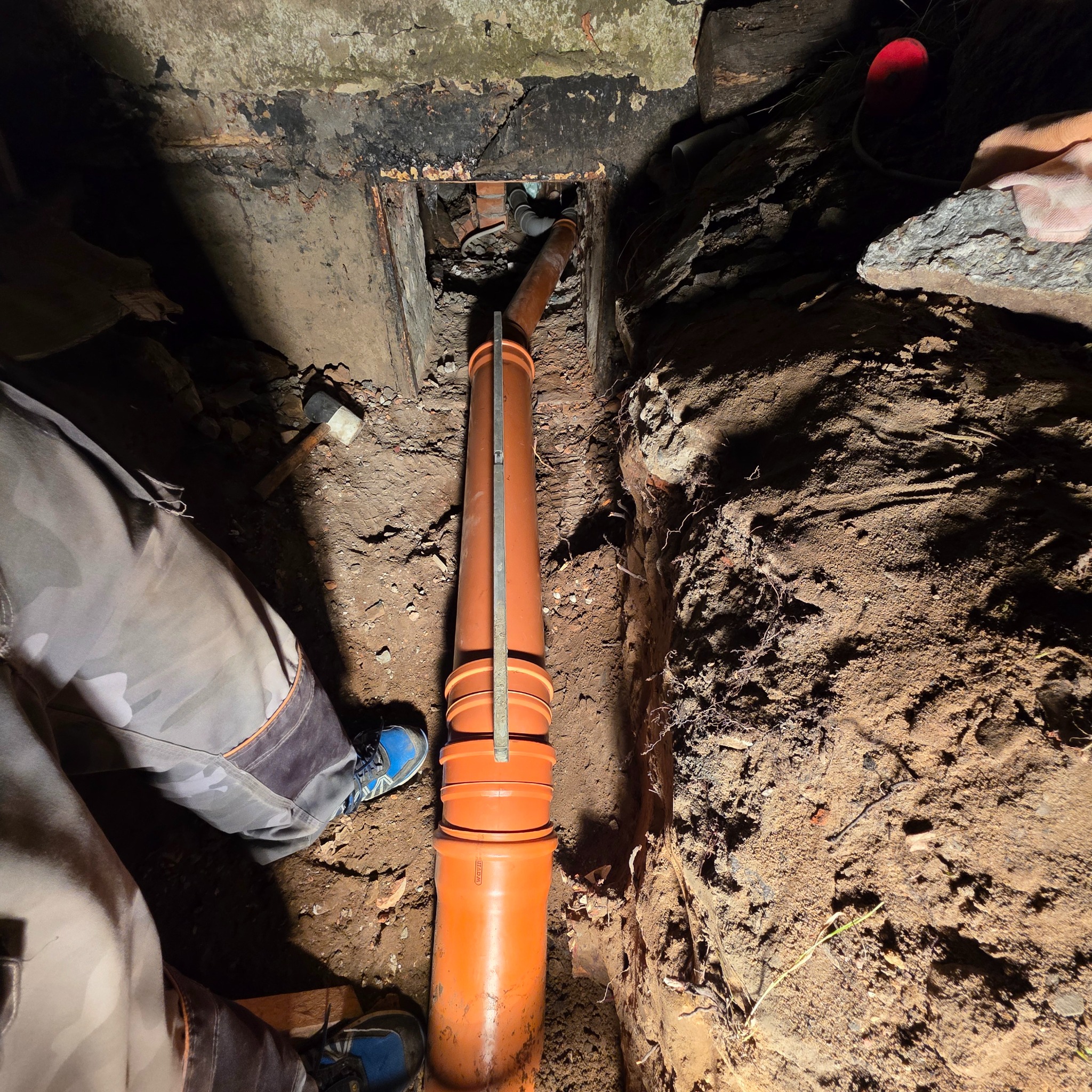 Pomarańczowa rura kanalizacyjna w wykopie ziemnym, widoczne połączenia. W tle fragment instalacji i ściana z cegieł. Prace ziemne i instalacyjne w trudnych warunkach.