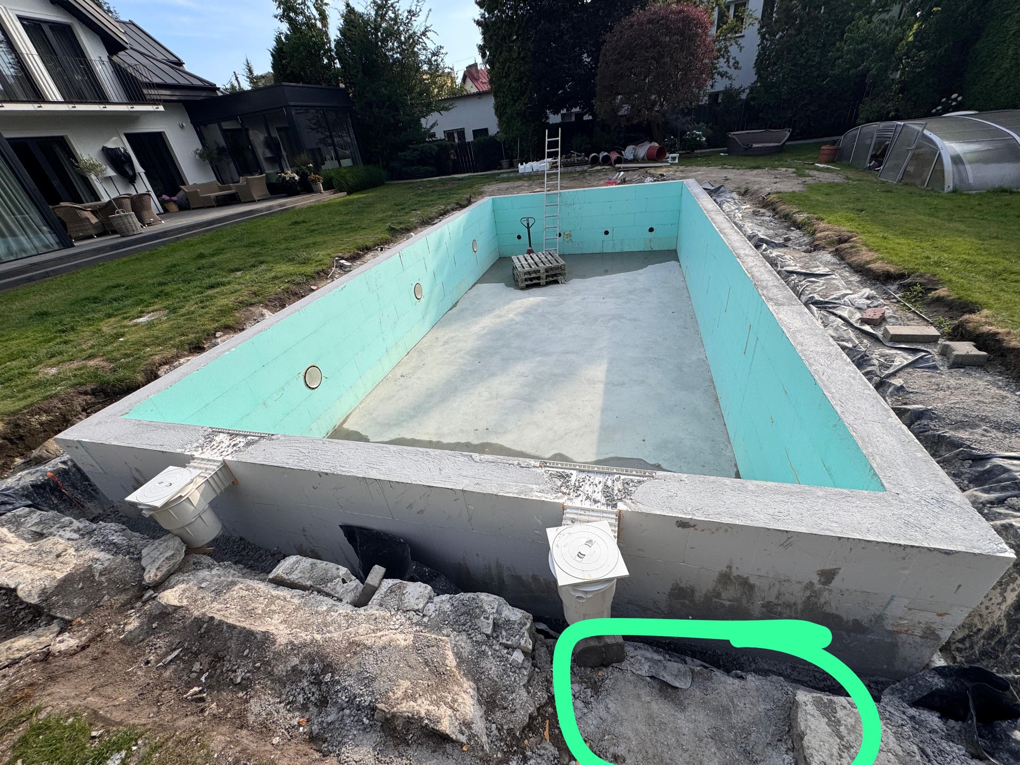 Budowa basenu w ogrodzie: betonowa konstrukcja z izolacją, widoczne prace ziemne i instalacje. Etap budowy, zieleń ogrodu w tle.