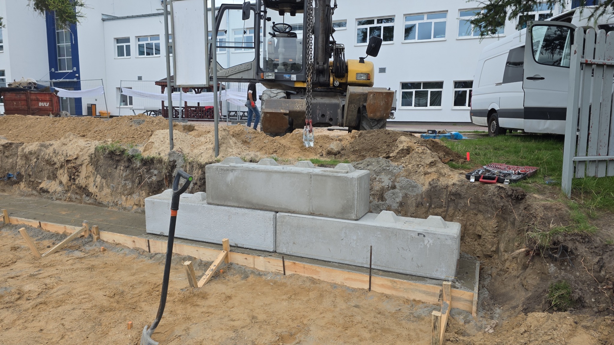 Montaż betonowych bloków na fundamencie z szalunkiem, koparka w tle. Widoczny wykop, narzędzia i budynek. Prace ziemne i fundamentowe w toku.