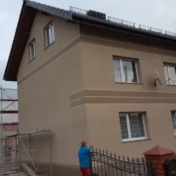 Dwupiętrowy dom z beżową elewacją z ciemniejszymi pasami, widoczne rusztowania i osoba w czerwonej czapce przy metalowym ogrodzeniu.