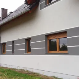 Ocieplony dom jednorodzinny z szarą elewacją w poziome pasy, brązowe okna z roletami, widok z boku na tle trawy.