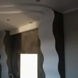 Wnętrze pokoju po remoncie z dekoracyjnymi, falistymi listwami ściennymi w kolorze białym i szarą ścianą pokrytą efektem betonu architektonicznego, oświetlenie punktowe w suficie.