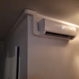 Biały klimatyzator ścienny marki Rotenso z wyświetlaczem pokazującym 20 stopni, z widocznymi białymi rurami instalacyjnymi, zamontowany w pomieszczeniu oświetlonym lampą sufitową.