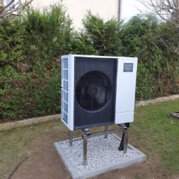 Biała pompa ciepła Mitsubishi Electric Zubadan R32, zamontowana na stalowej konstrukcji na betonowej podstawie z białymi kamykami, na trawniku przed żywopłotem.