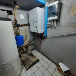 Instalacja kotła gazowego Ecodan Mitsubishi Electric z podłączeniem do zasobnika ciepłej wody i niebieskiego naczynia przeponowego w pomieszczeniu technicznym z szarą ścianą i białą glazurą podłogową.