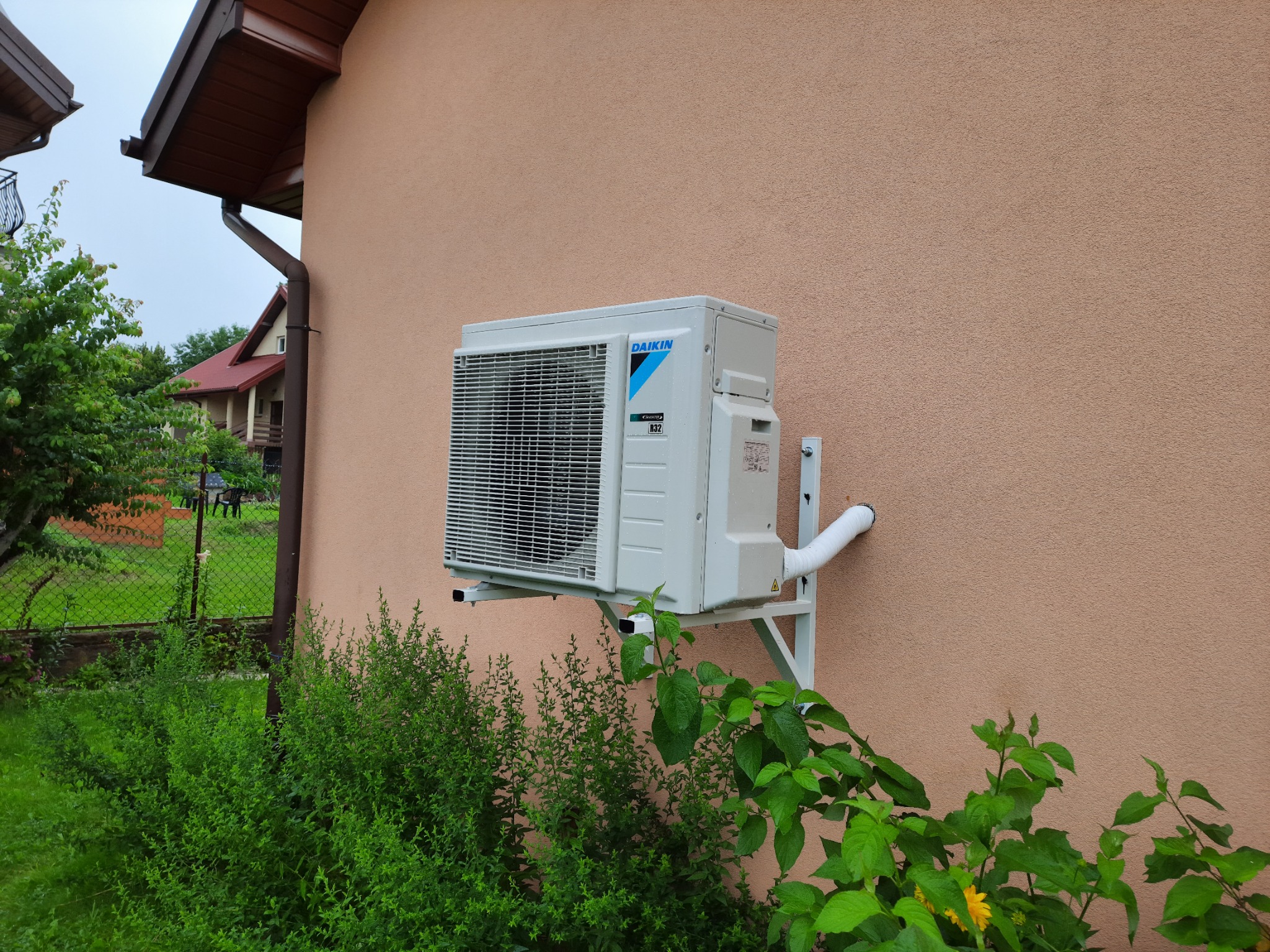 Zewnętrzna jednostka klimatyzacji Daikin zamontowana na ścianie domu z beżowym tynkiem, widoczny biały odpływ skroplin i fragment ogrodu z zielenią.