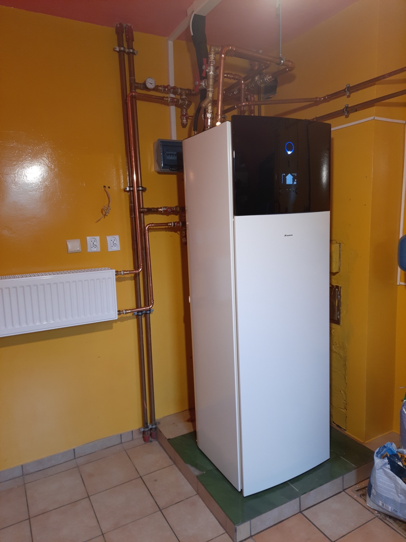 Zainstalowana biało-czarna pompa ciepła Daikin w pomieszczeniu z żółtymi ścianami, widoczne miedziane rury instalacji grzewczej oraz grzejnik.