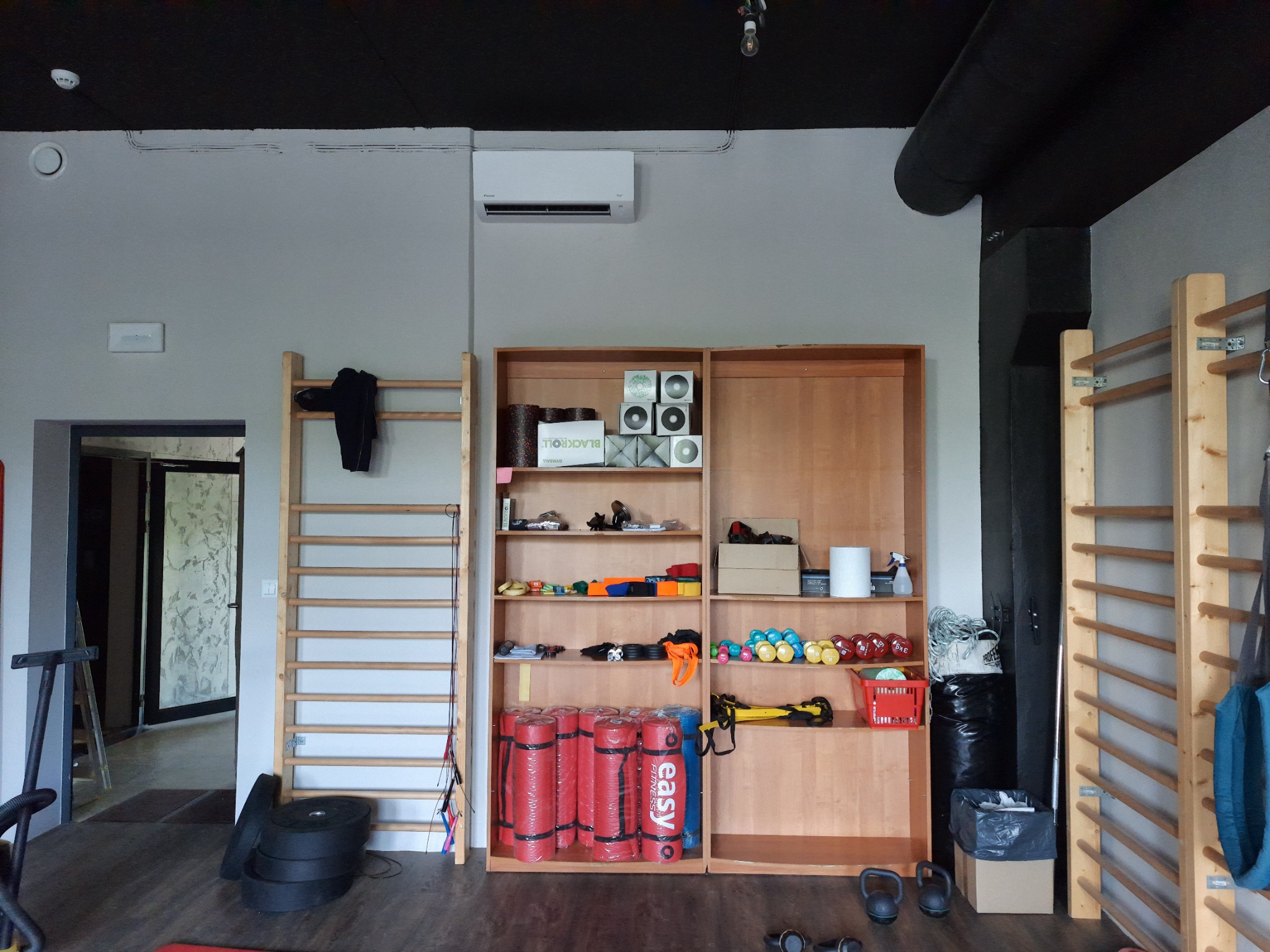 Wnętrze siłowni z klimatyzatorem na ścianie, drewnianymi drabinkami gimnastycznymi, regałem z akcesoriami do ćwiczeń, takimi jak maty, ciężarki i piłki.