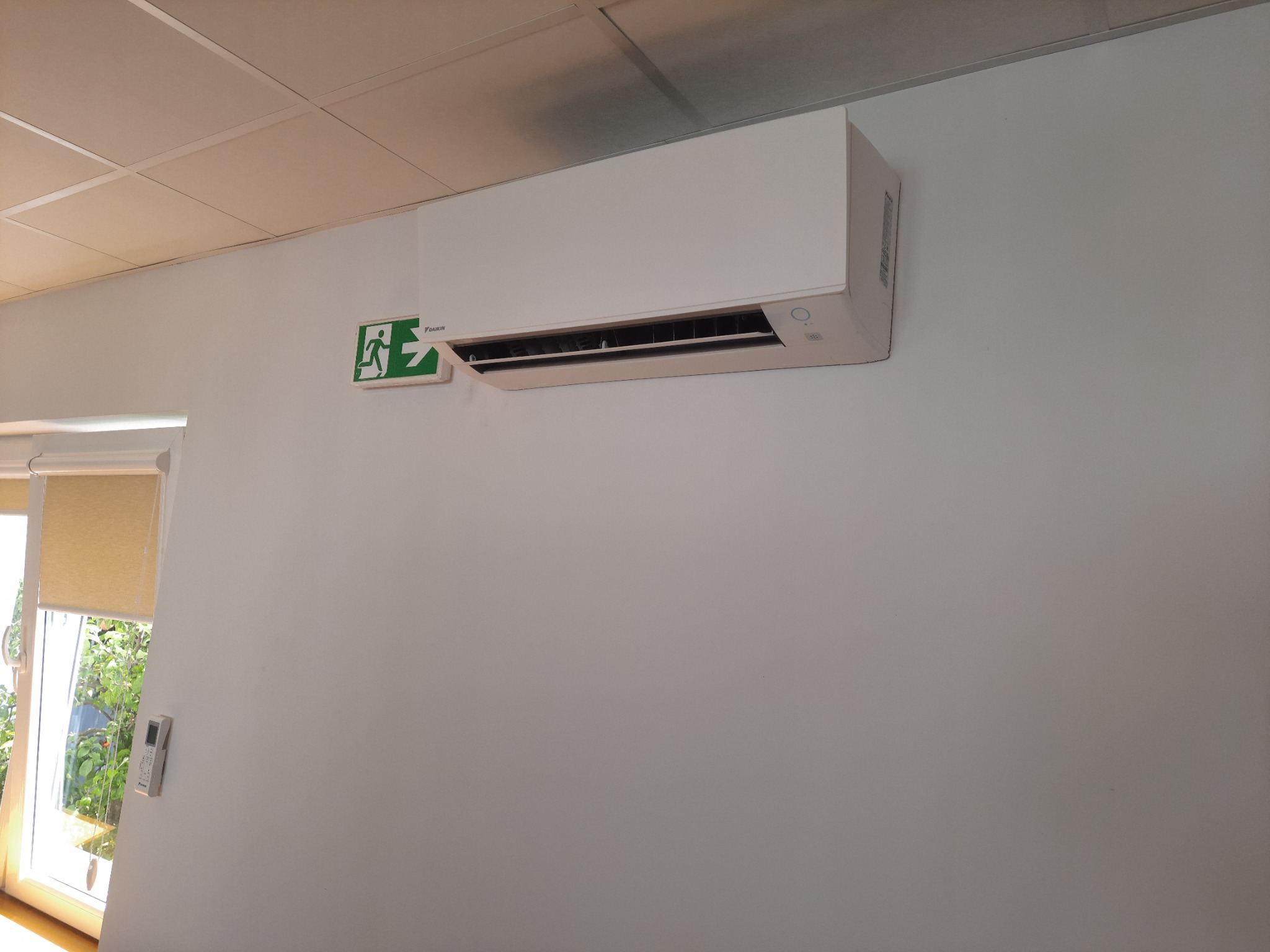 Biała jednostka klimatyzacji Daikin zamontowana na ścianie biura, obok zielonego znaku ewakuacyjnego. W tle widoczne okno z roletą i zielenią.