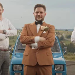 Pan młody w brązowym garniturze i muszce, pozuje z dwoma mężczyznami przy błękitnym Fiacie 126p na tle pagórkowatego krajobrazu.