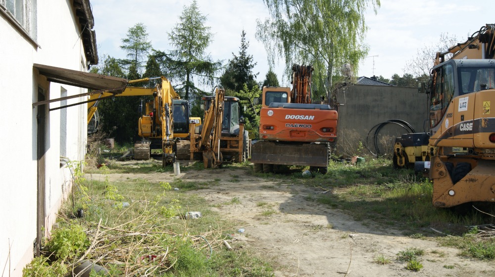 Kilka maszyn budowlanych, w tym pomarańczowa koparka DOOSAN, stoją zaparkowane na placu obok budynku, widoczne także żółte koparki i walec.