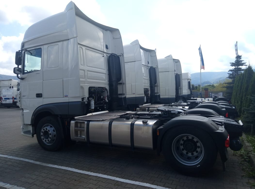 Rząd białych ciągników siodłowych DAF zaparkowanych na placu, widok z boku, w tle flaga i zalesione wzgórza.