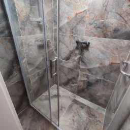 Szklana kabina prysznicowa z armaturą i odpływem liniowym, wykończona płytkami imitującymi marmur w odcieniach szarości i brązu.