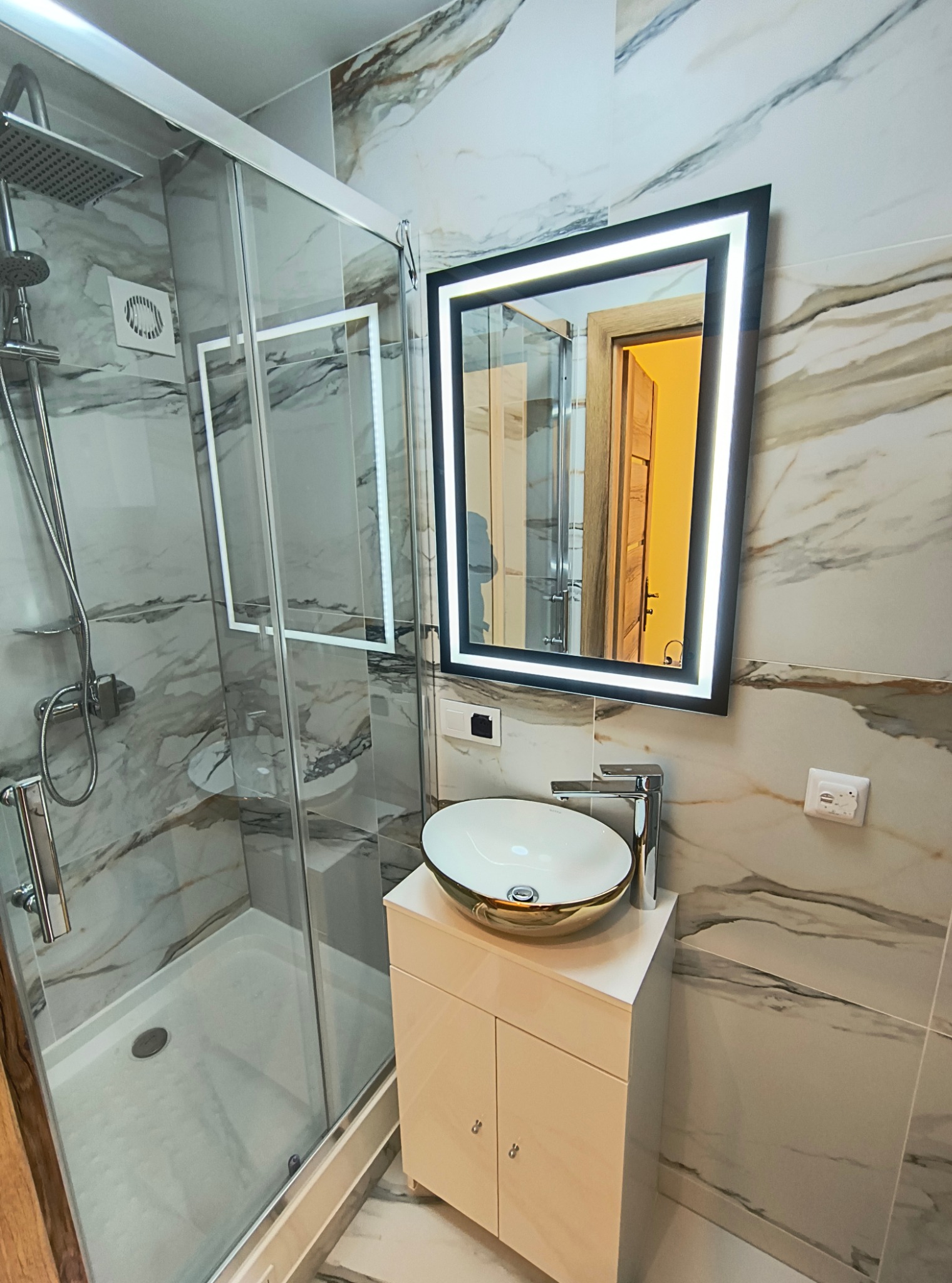 Nowoczesna łazienka z kabiną prysznicową, umywalką nablatową w kolorze białym ze złotym rantem, podświetlanym lustrem w czarnej ramie i marmurowymi płytkami.