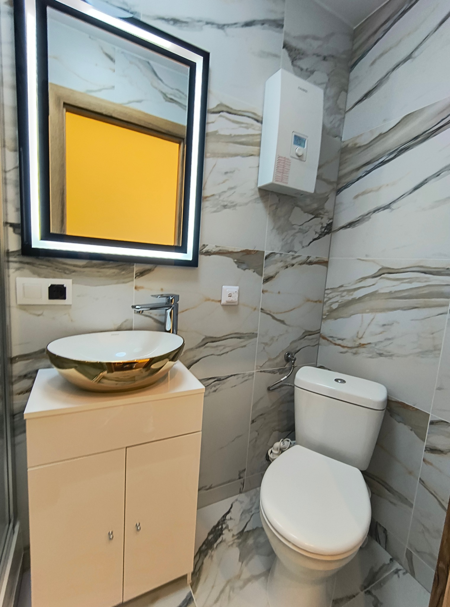 Wykończona łazienka z marmurowymi płytkami, złotą umywalką nablatową i podświetlanym lustrem LED, widoczny bojler elektryczny.