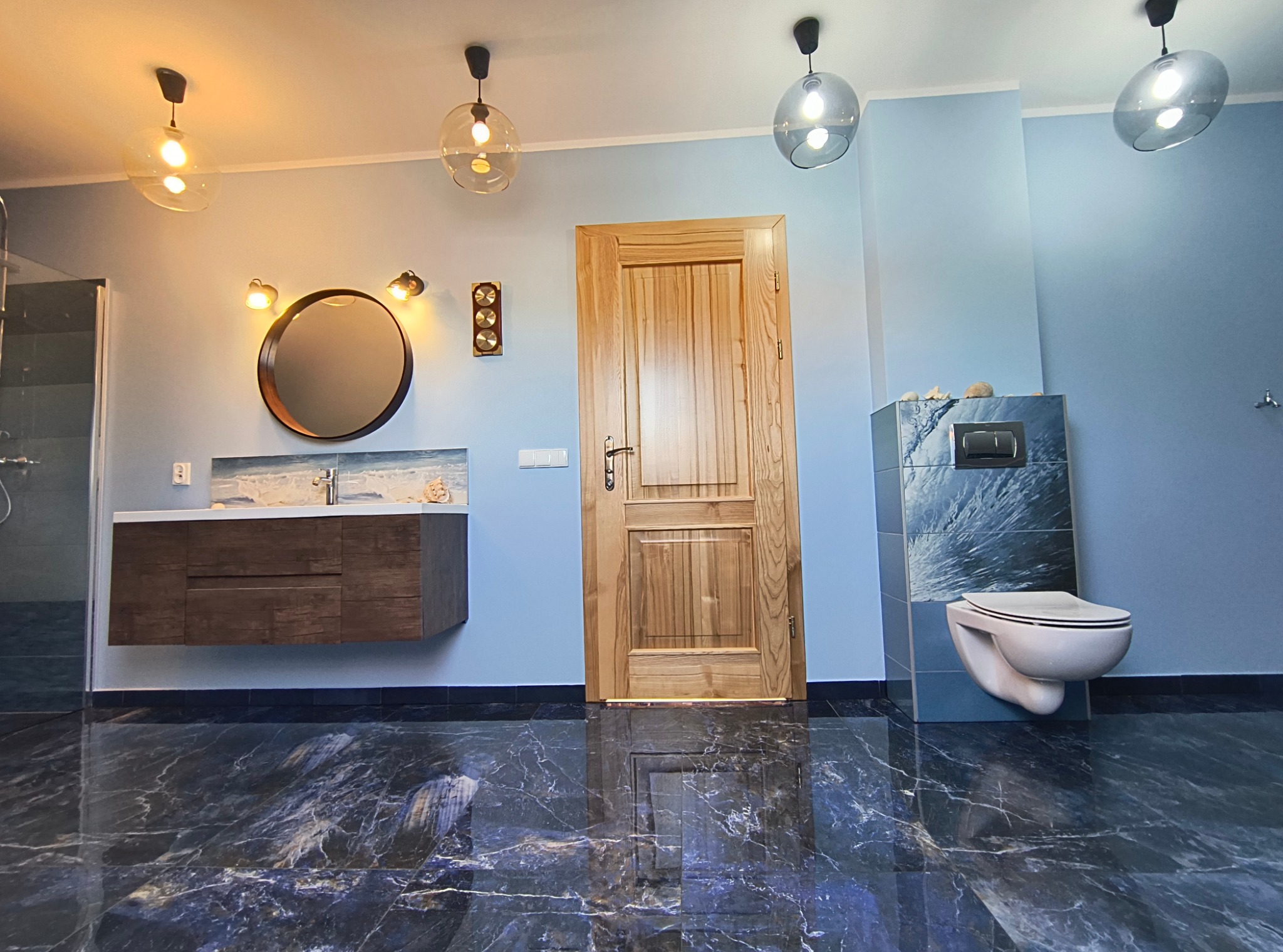 Wyremontowana łazienka z drewnianą umywalką, okrągłym lustrem z oświetleniem, podwieszaną toaletą z niebieską mozaiką i drewnianymi drzwiami, wykończona na niebiesko, z podłogą z ciemnego marmuru.