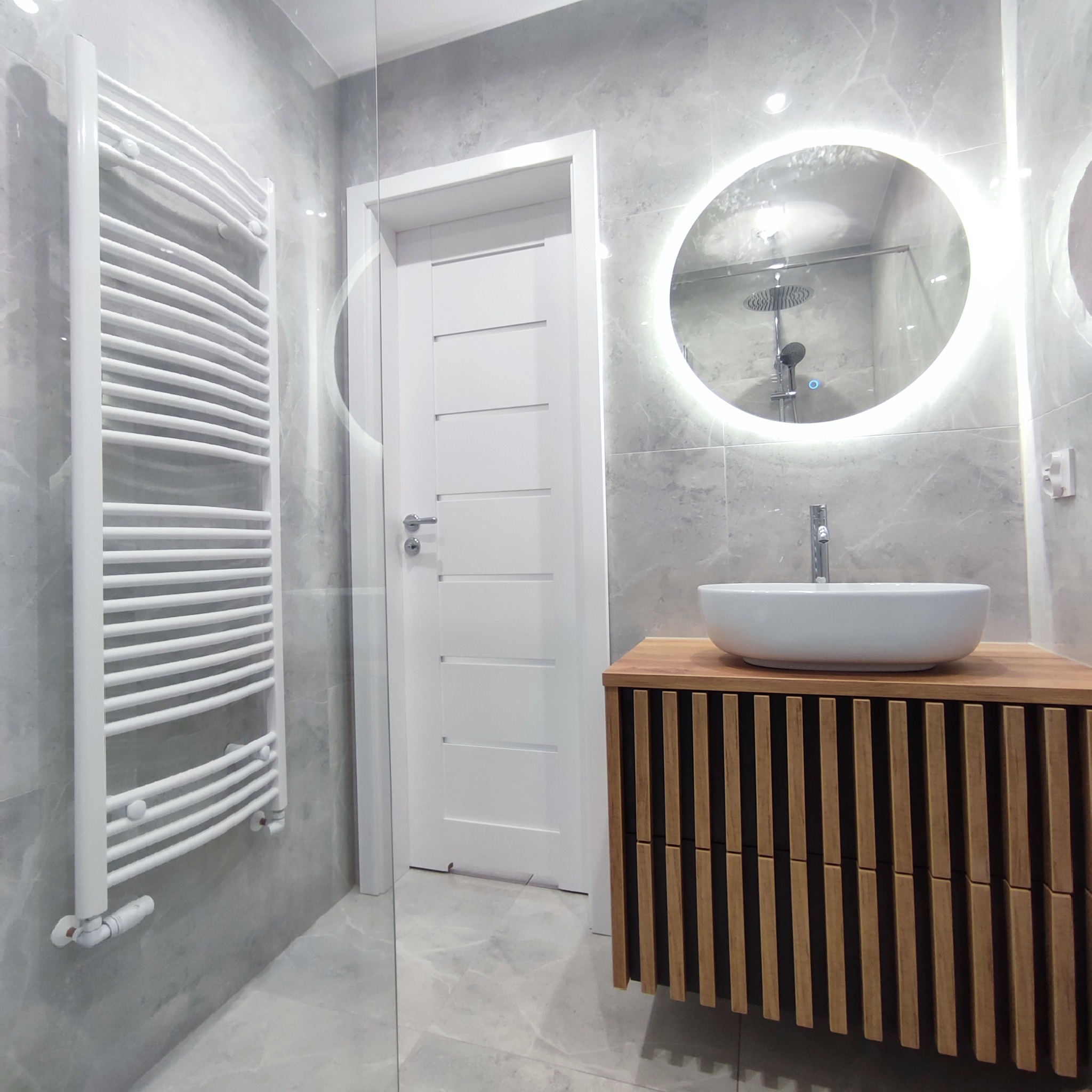 Nowoczesna łazienka z szarymi marmurowymi płytkami, podświetlanym okrągłym lustrem, białą umywalką nablatową na drewnianej szafce z lamelami, białym grzejnikiem drabinkowym i białymi drzwiami.