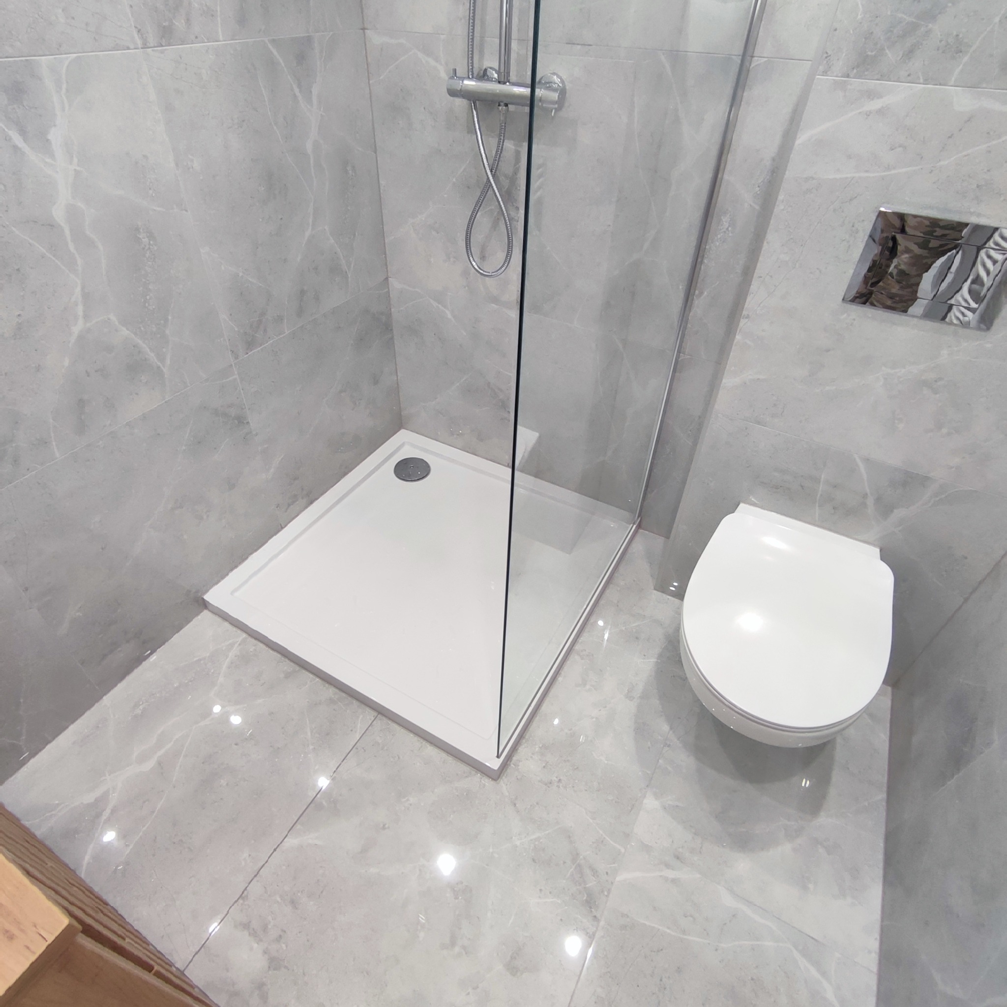 Nowoczesna łazienka wykończona szarymi płytkami imitującymi marmur, z kabiną prysznicową typu walk-in z białą brodzikiem i wiszącą toaletą.