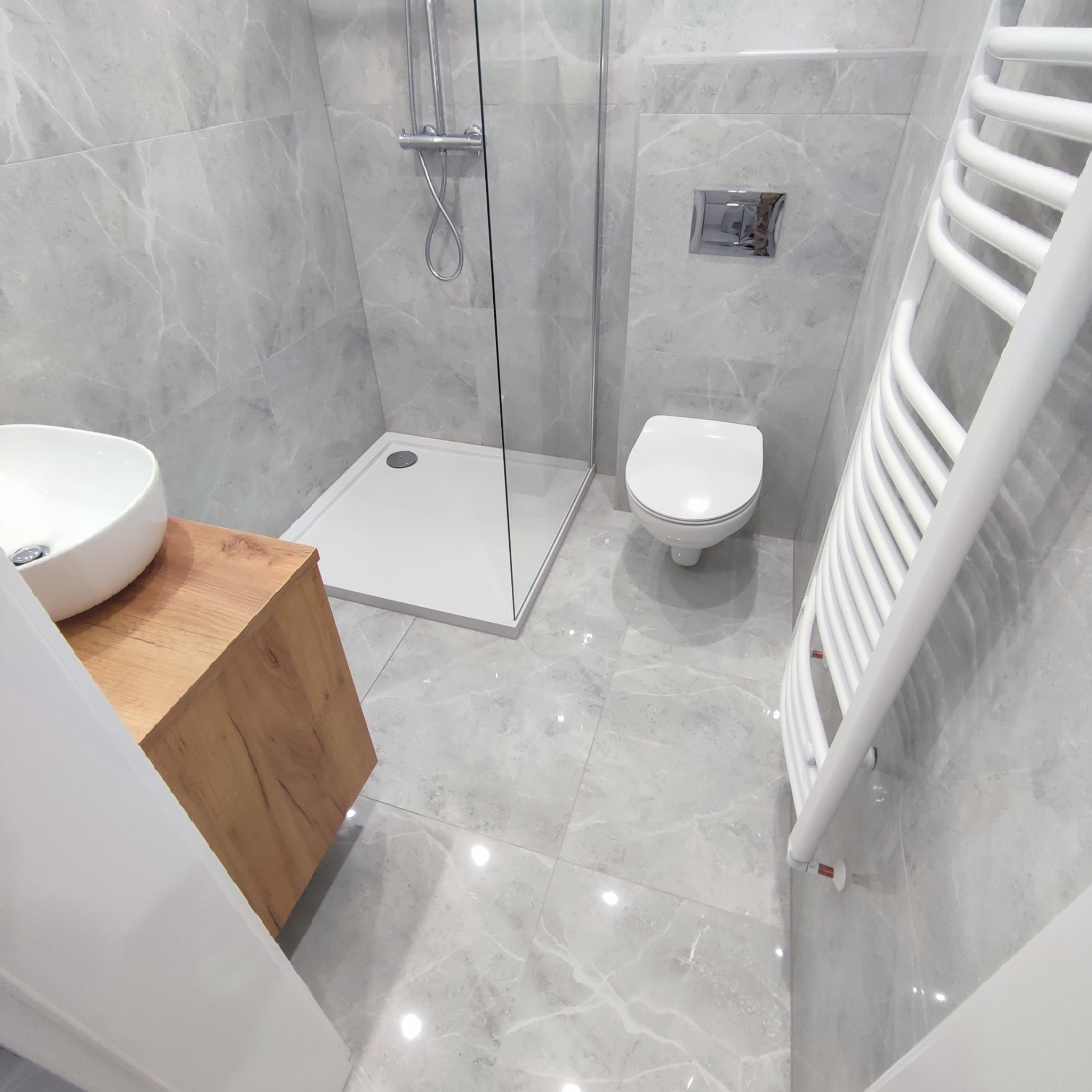 Nowoczesna łazienka z szarymi marmurkowymi płytkami, kabiną prysznicową ze szklaną ścianką, umywalką nablatową na drewnianej szafce i białym grzejnikiem drabinkowym.