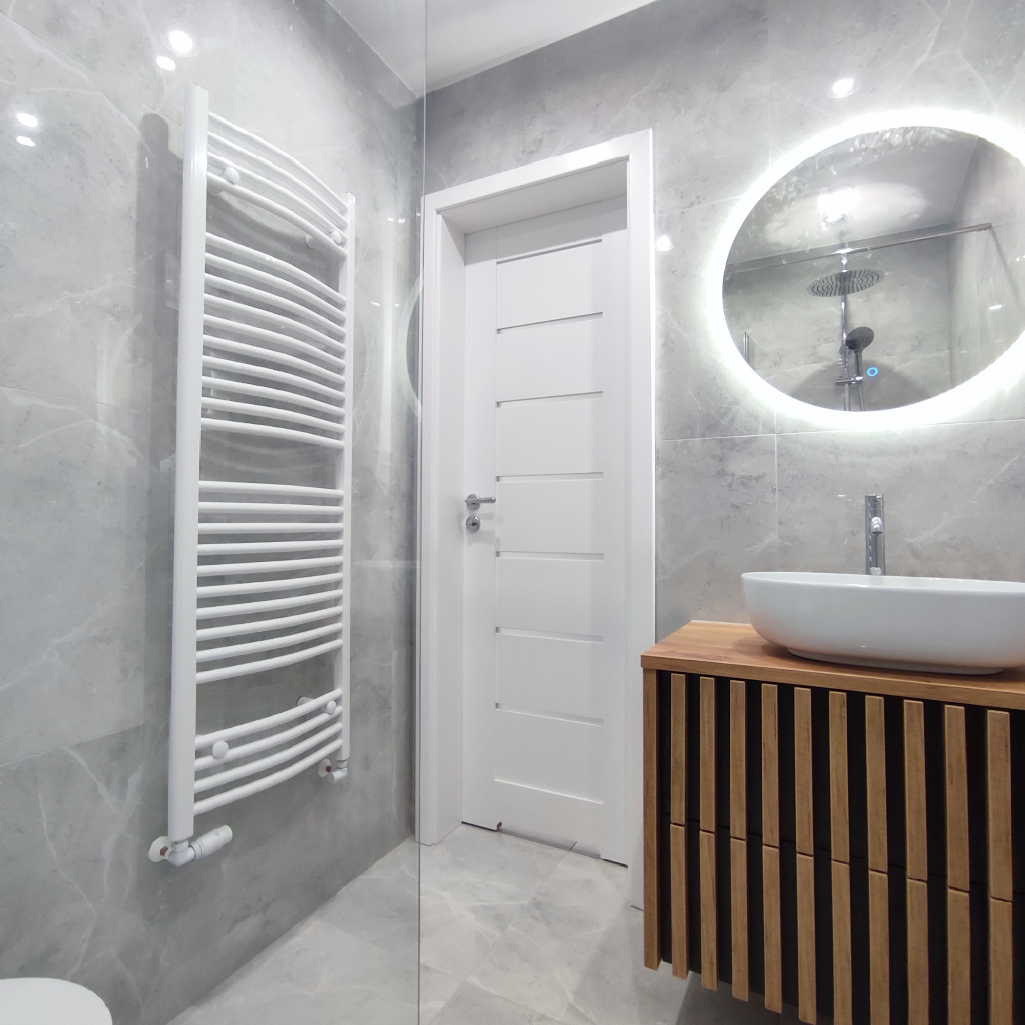 Nowoczesna łazienka z szarymi marmurowymi płytkami, białym grzejnikiem drabinkowym, okrągłym lustrem z podświetleniem LED, umywalką nablatową na drewnianej szafce z lamelami i białą ścianą z drzwiami.