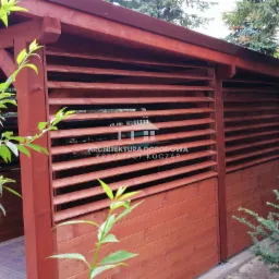 Czerwono-brązowa, drewniana konstrukcja z poziomymi lamelami, otoczona zielenią, z widocznym fragmentem brukowanej ścieżki wewnątrz.