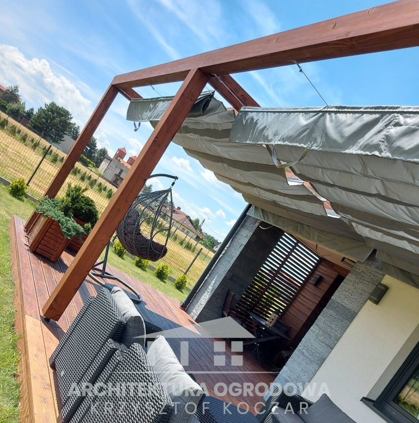Drewniana konstrukcja z rozsuwanym zadaszeniem nad tarasem z meblami ogrodowymi i wiszącym fotelem, widok z boku, w tle ogrodzenie i zabudowa jednorodzinna.