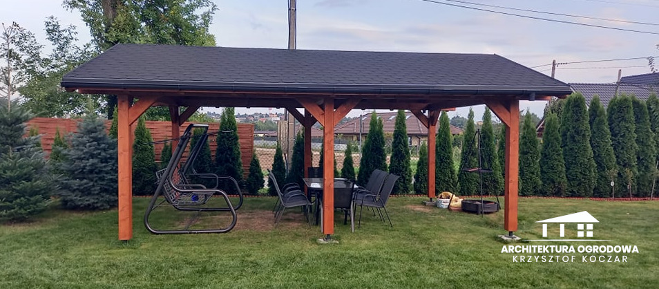 Zadaszona konstrukcja z drewna z czarnym dachem, wyposażona w stół, krzesła i huśtawkę ogrodową, otoczona zielonym trawnikiem i tujami.