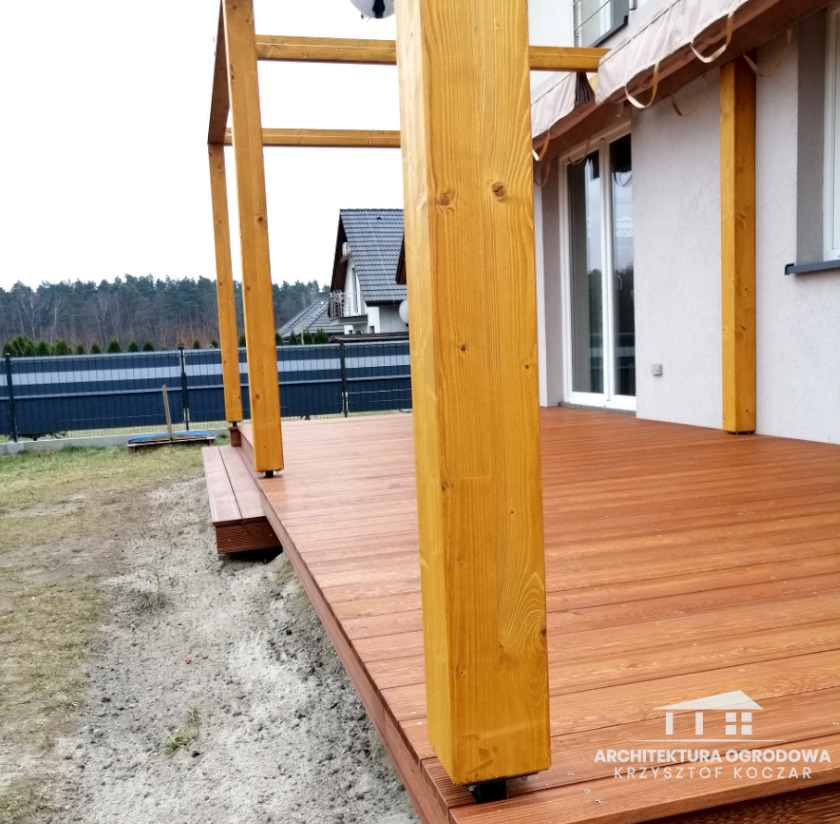 Drewniany taras z jasnymi, pionowymi słupami konstrukcyjnymi, widok z boku, z logo firmy w prawym dolnym rogu.