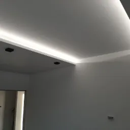 Nowoczesne oświetlenie sufitowe LED w kształcie litery L, z czarnymi punktowymi lampami, świeżo pomalowane białe ściany i widoczne drzwi do innego pomieszczenia.