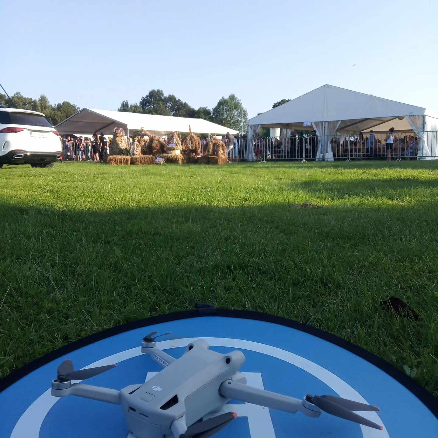 Szary dron DJI Mini 3 Pro na niebieskiej macie startowej, w tle zielony trawnik, białe namioty z ludźmi i biały samochód, prawdopodobnie podczas imprezy plenerowej.