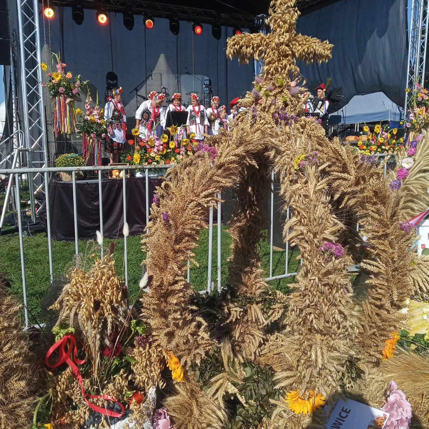 Wieniec dożynkowy w kształcie krzyża i korony, wykonany ze zboża i kwiatów, na tle sceny z zespołem ludowym w tradycyjnych strojach.