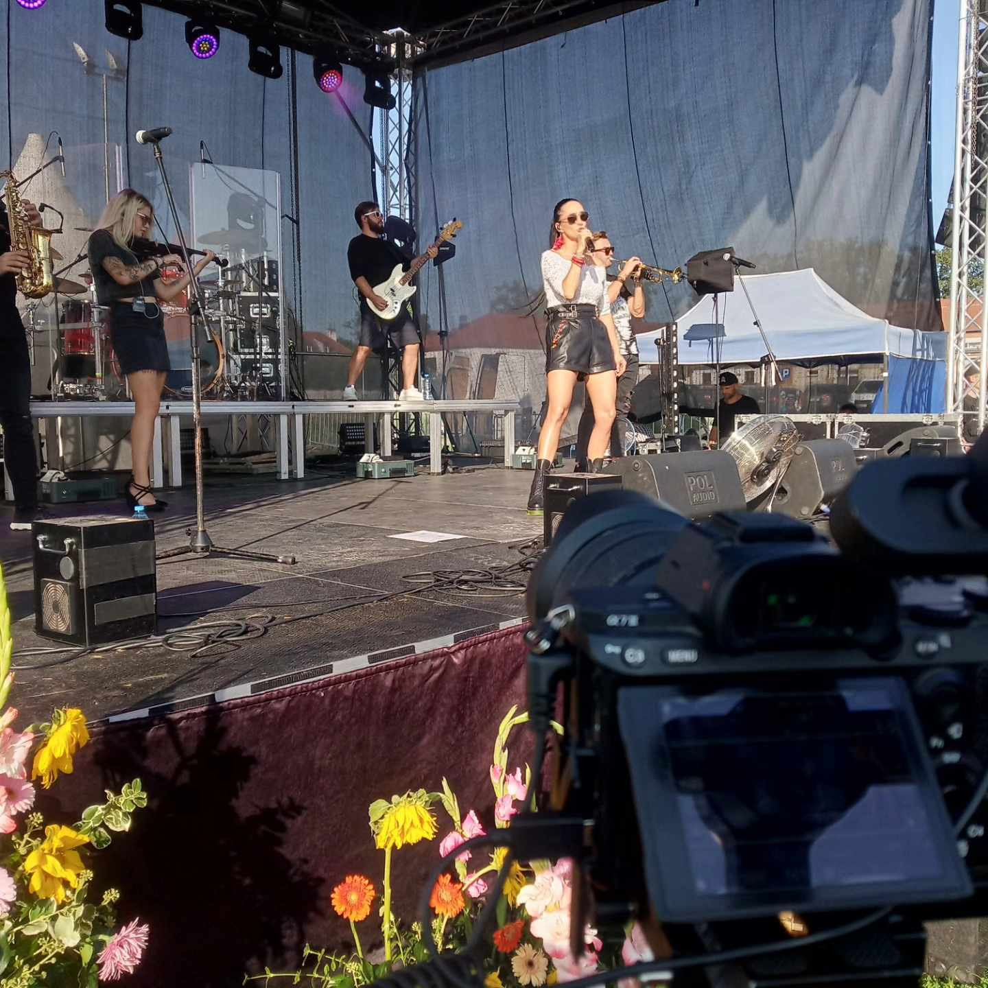 Kamera filmowa rejestruje występ zespołu muzycznego na scenie plenerowej, wokalistka w skórzanych szortach i trębacz w tle, z perspektywy operatora.