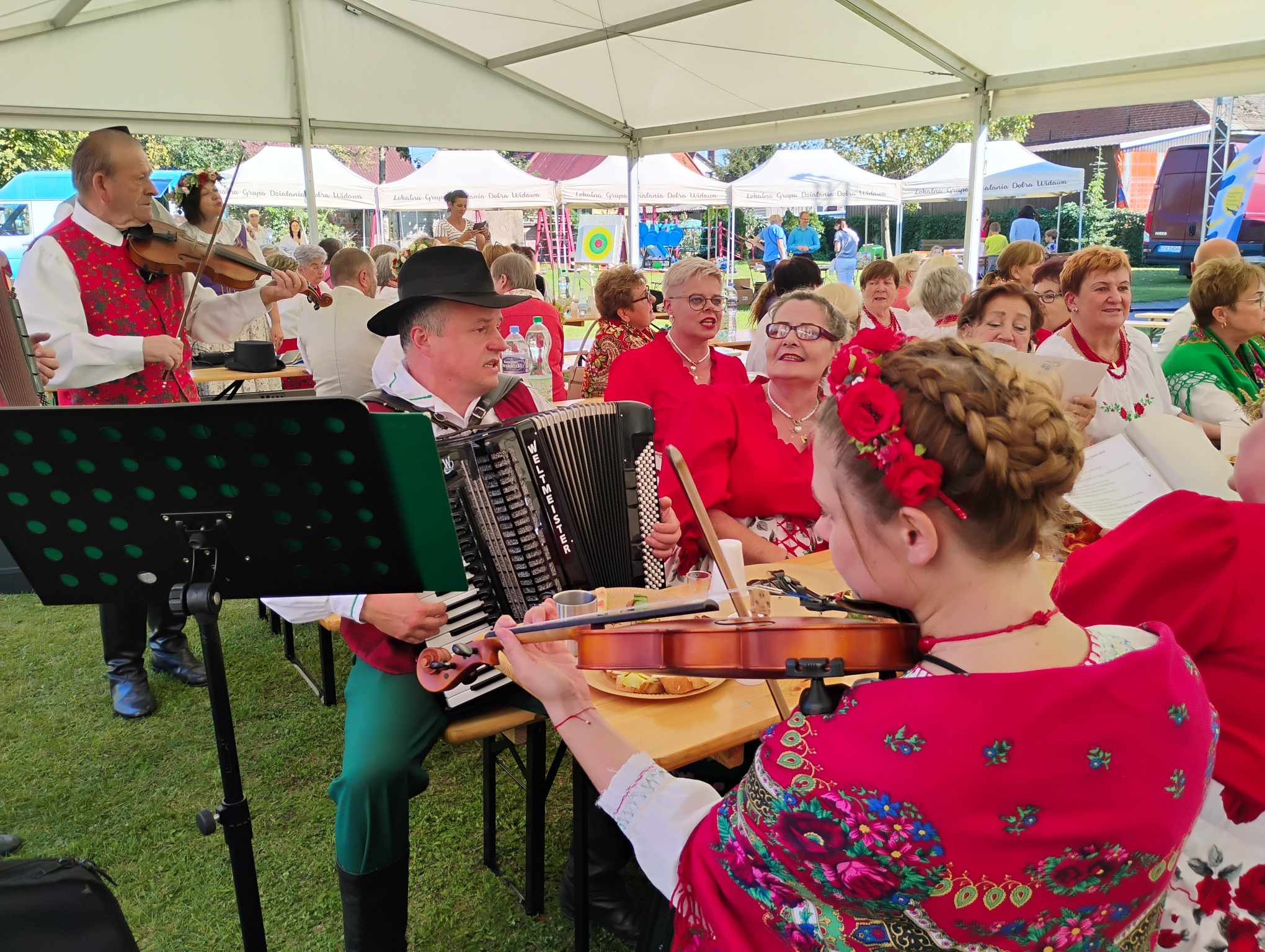 Zespół muzyczny w strojach ludowych grający na akordeonie i skrzypcach podczas plenerowej imprezy, widownia w tle.