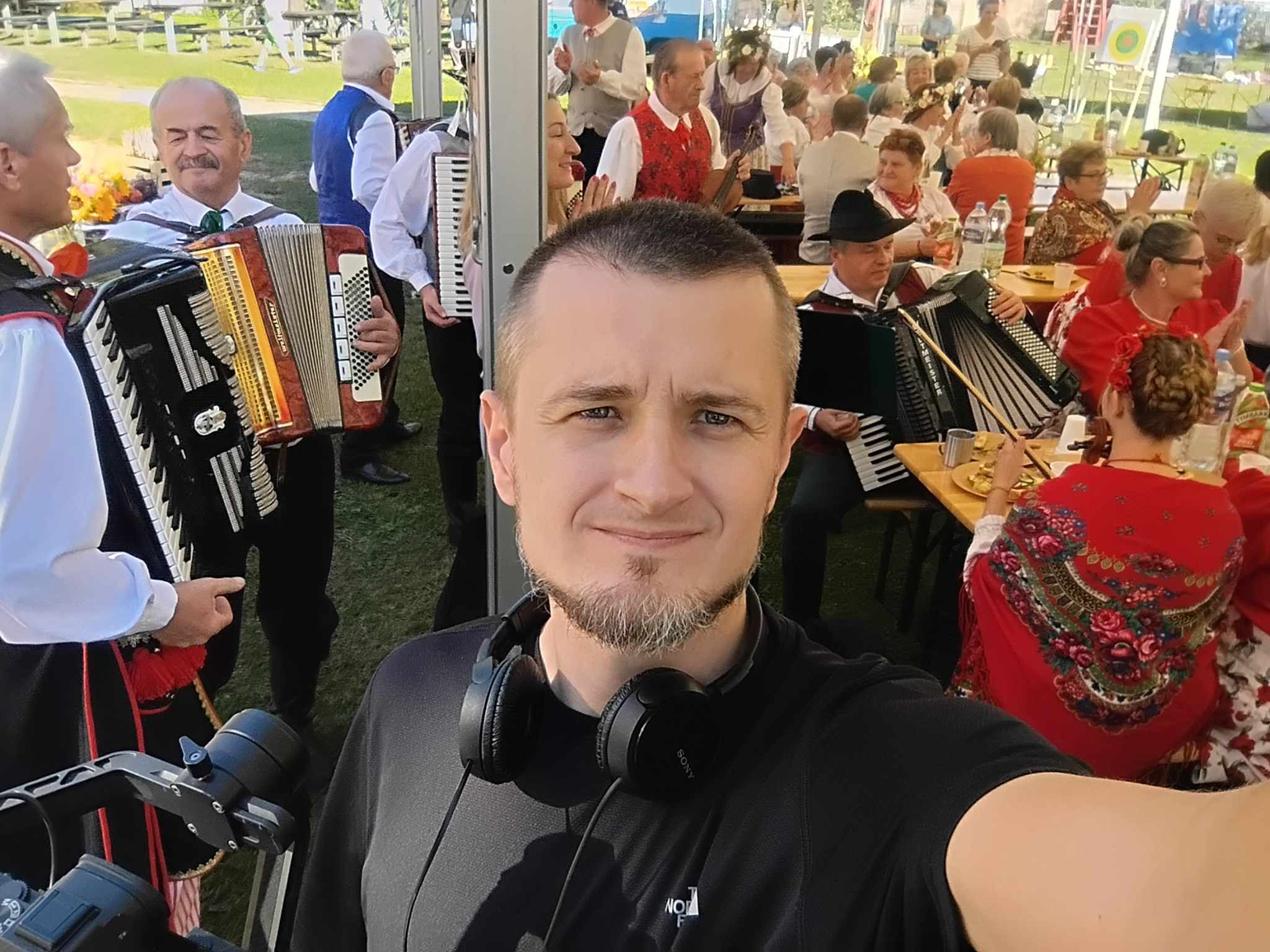 Selfie mężczyzny z brodą i słuchawkami na szyi na tle występu akordeonistów w strojach ludowych podczas plenerowej imprezy.