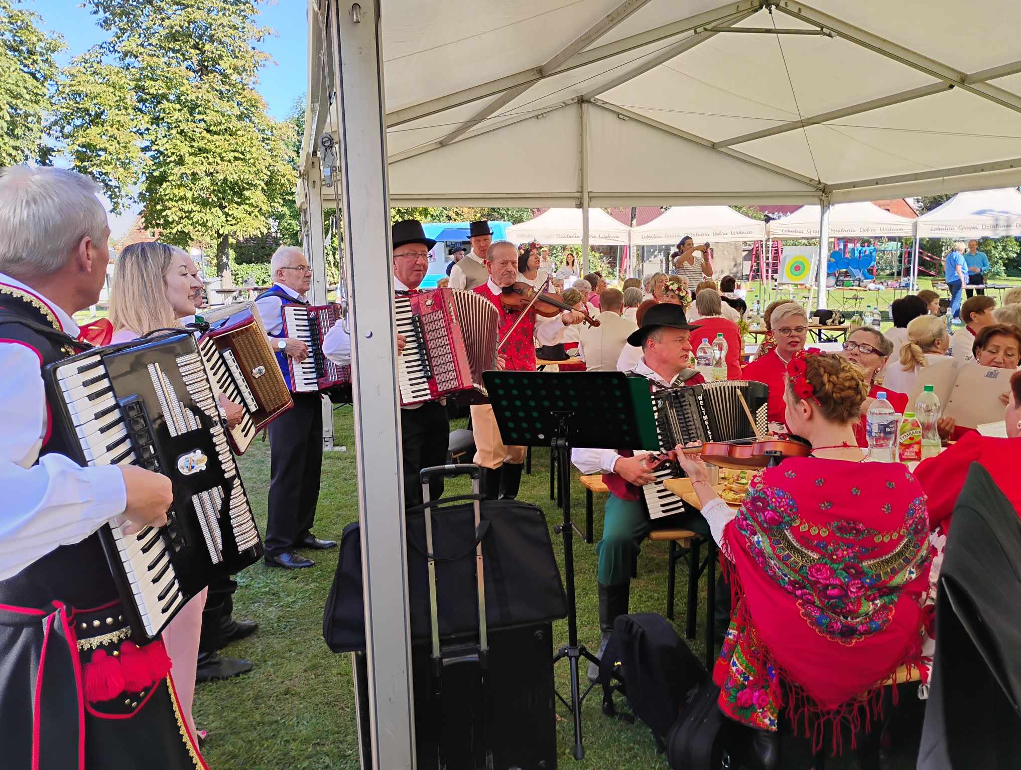 Zespół muzyczny w strojach ludowych grający na akordeonach i skrzypcach pod namiotem podczas plenerowej imprezy.