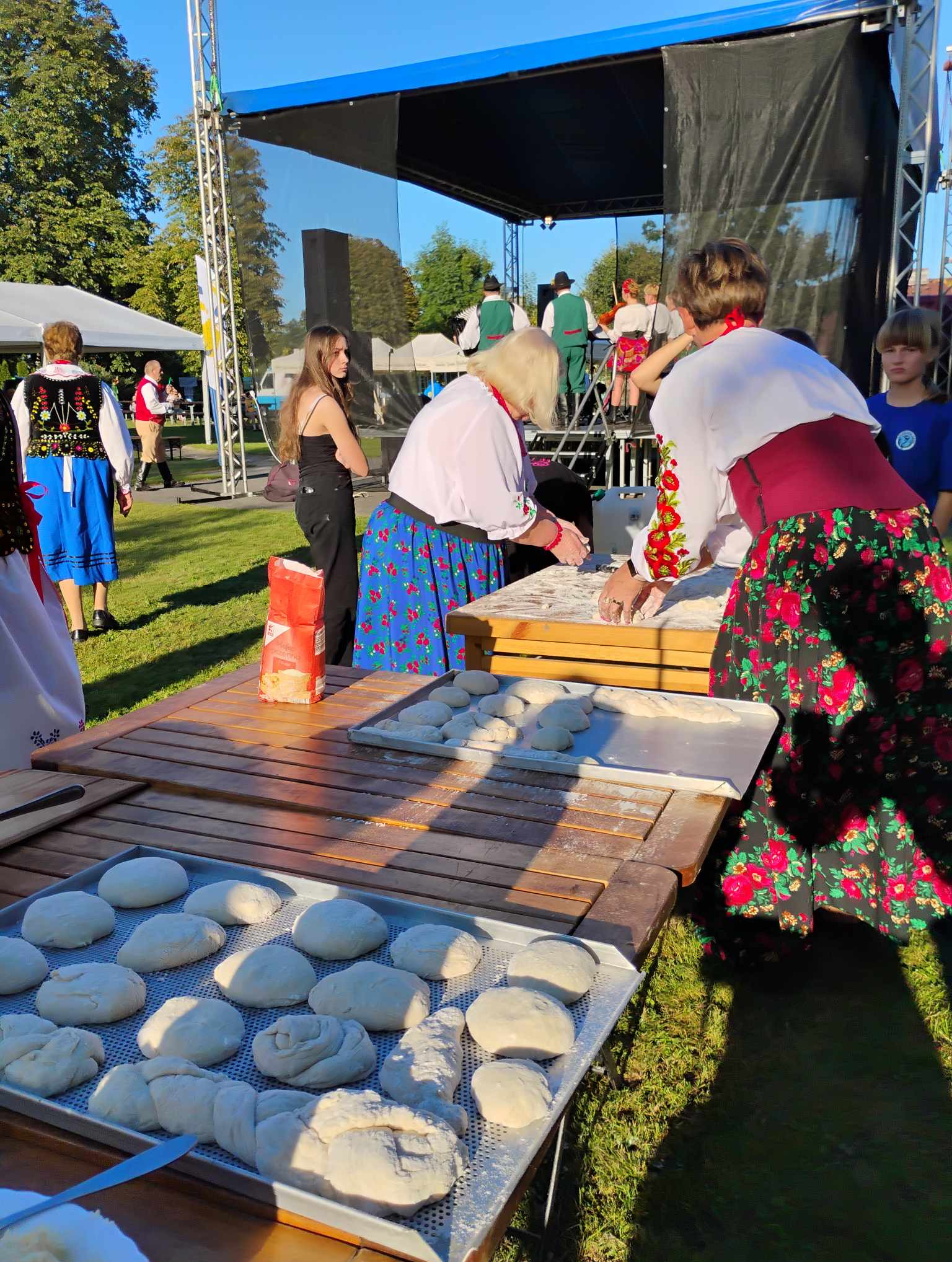 Przygotowywanie ciasta na festynie ludowym: kobiety w strojach regionalnych formują bułki na blasze, w tle scena z występującymi muzykami, na stole mąka.