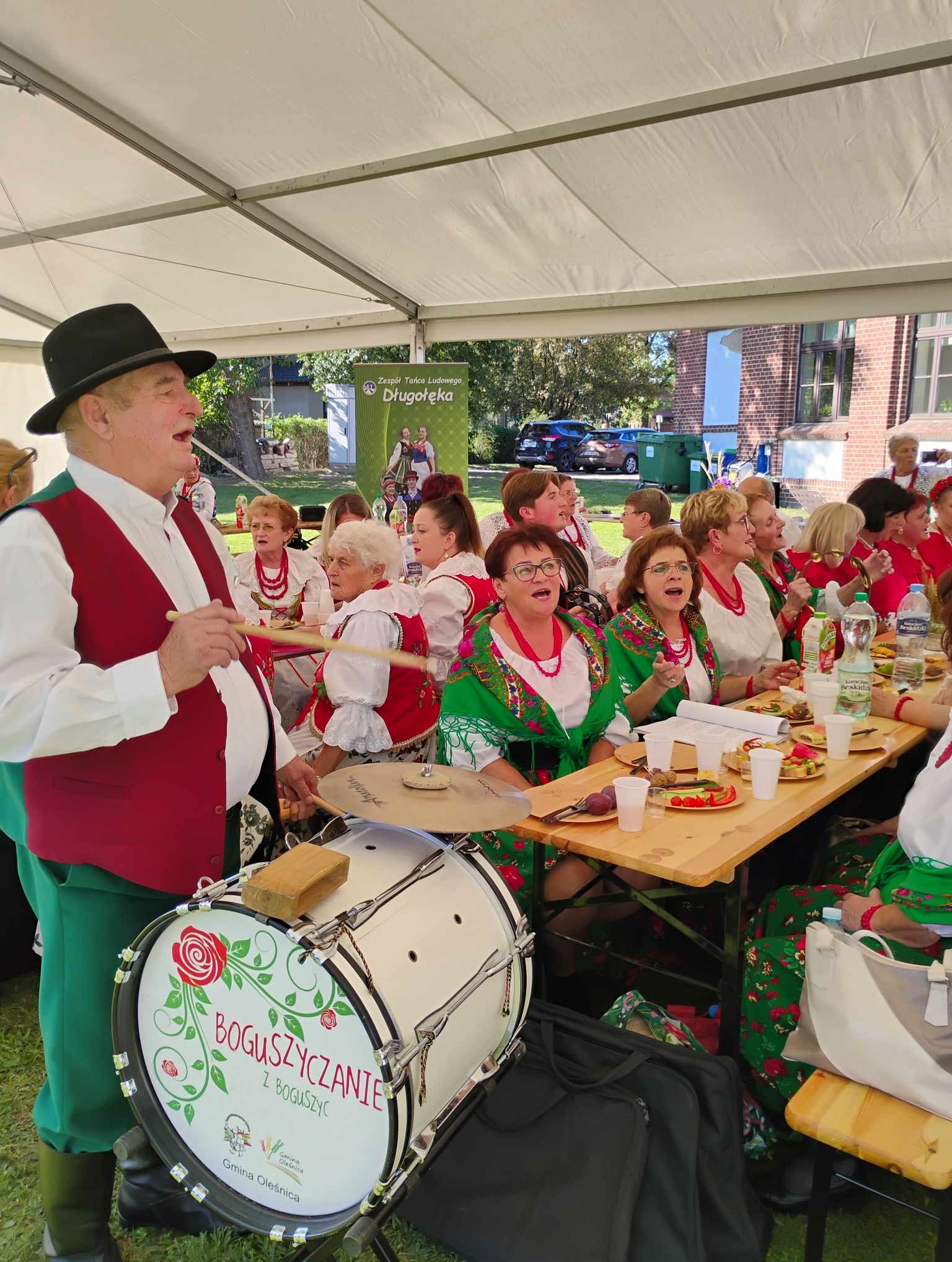 Zespół ludowy 'Boguszyczanie' podczas występu: mężczyzna w stroju ludowym gra na bębnie, a kobiety w tradycyjnych strojach śpiewają przy stole, pod namiotem.