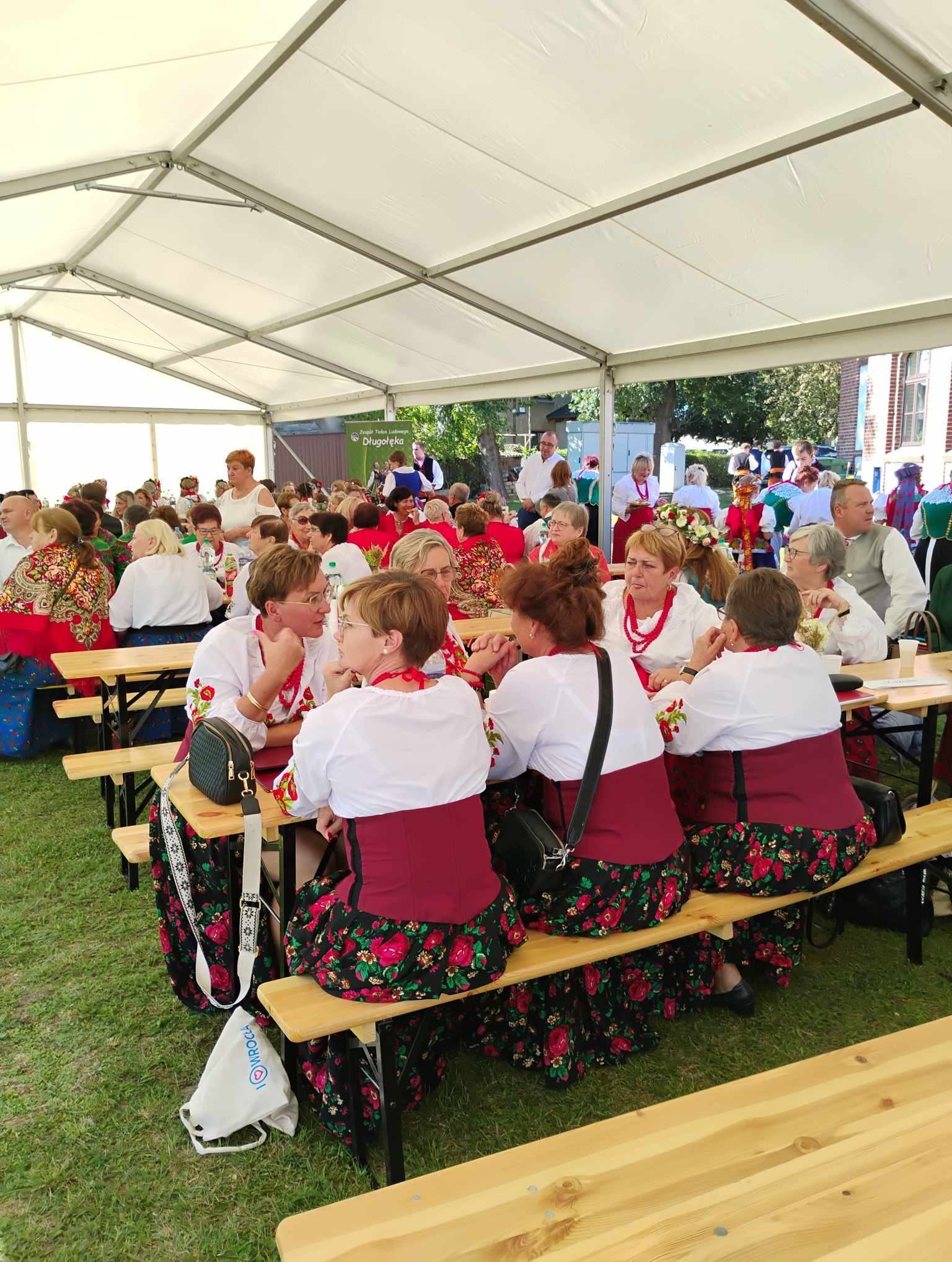 Grupa kobiet w strojach ludowych siedząca przy drewnianych stołach pod białym namiotem, widoczne detale haftu na bluzkach i wzory na spódnicach, w tle inne osoby i budynki.