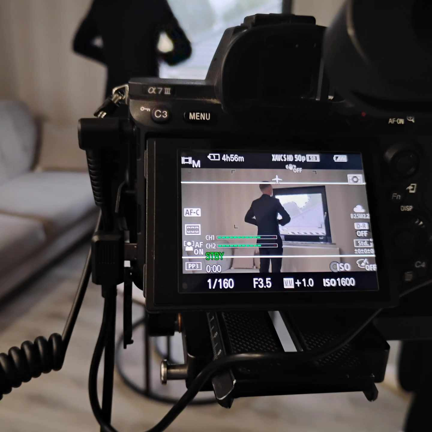 Ekran aparatu Sony Alpha 7 III podczas nagrywania wideo z mężczyzną w garniturze na tle jasnego pomieszczenia, widoczne ustawienia ekspozycji i parametry nagrywania.