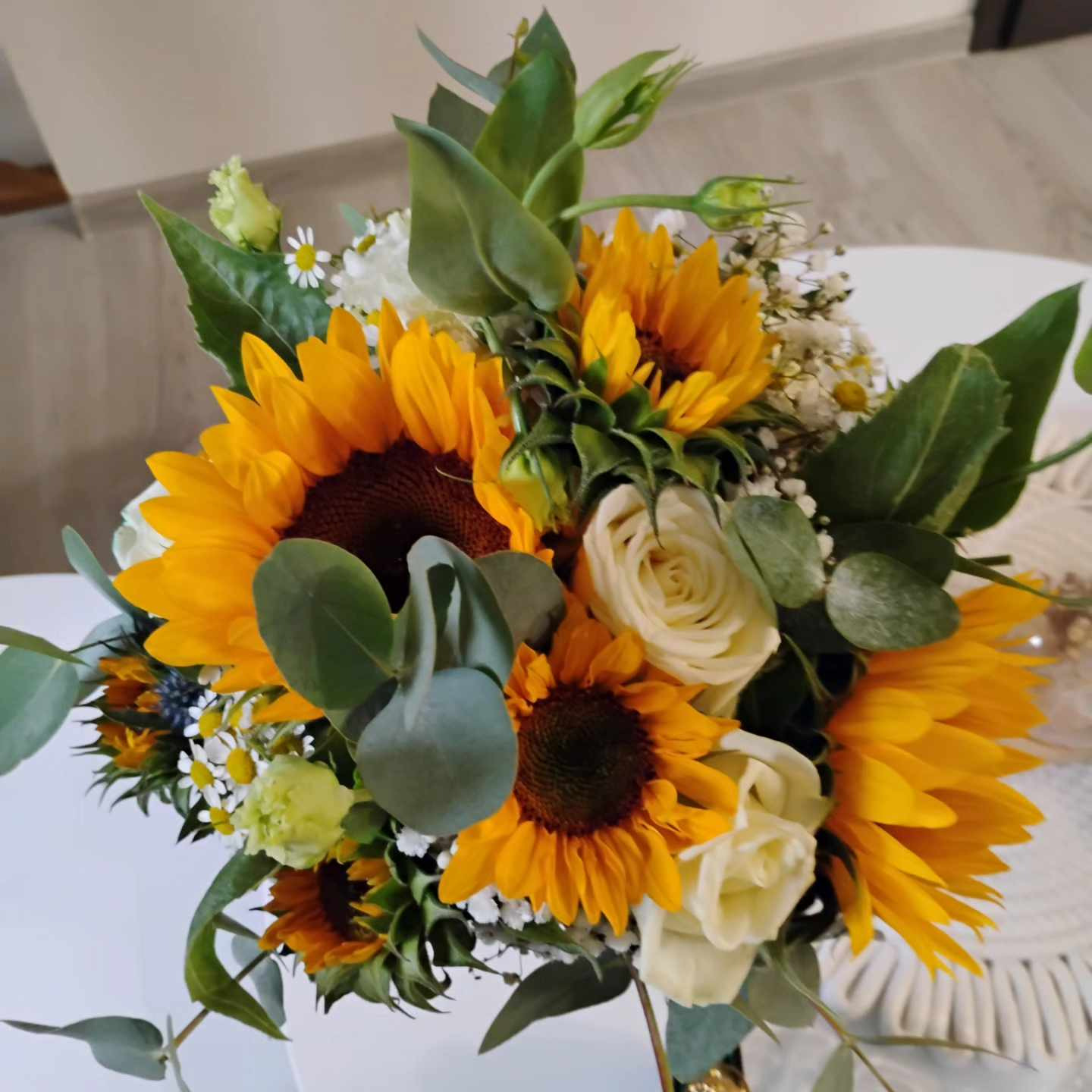 Bukiet ślubny z przewagą słoneczników, białych róż i zielonych liści eukaliptusa, ułożony na białym stole z elementami dekoracji makramy w tle.