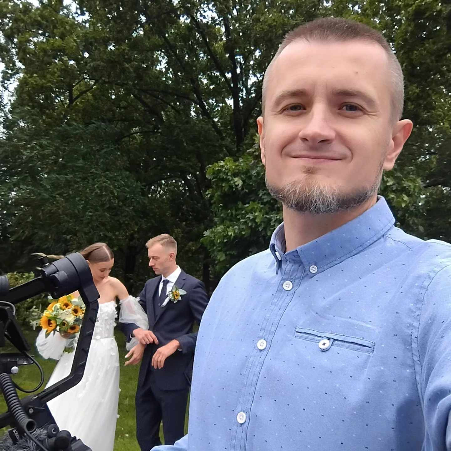 Uśmiechnięty mężczyzna w jasnoniebieskiej koszuli, w tle para młoda i kamera na statywie podczas sesji plenerowej.