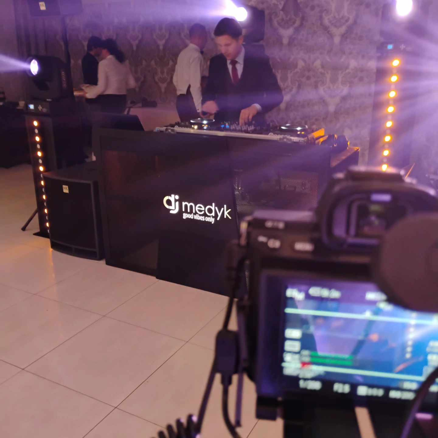 Kamerzysta filmuje DJ-a w garniturze, który miksuje muzykę na konsolecie z logo 'dj medyk good vibes only'. W tle oświetlenie sceniczne i dekoracyjna tapeta.