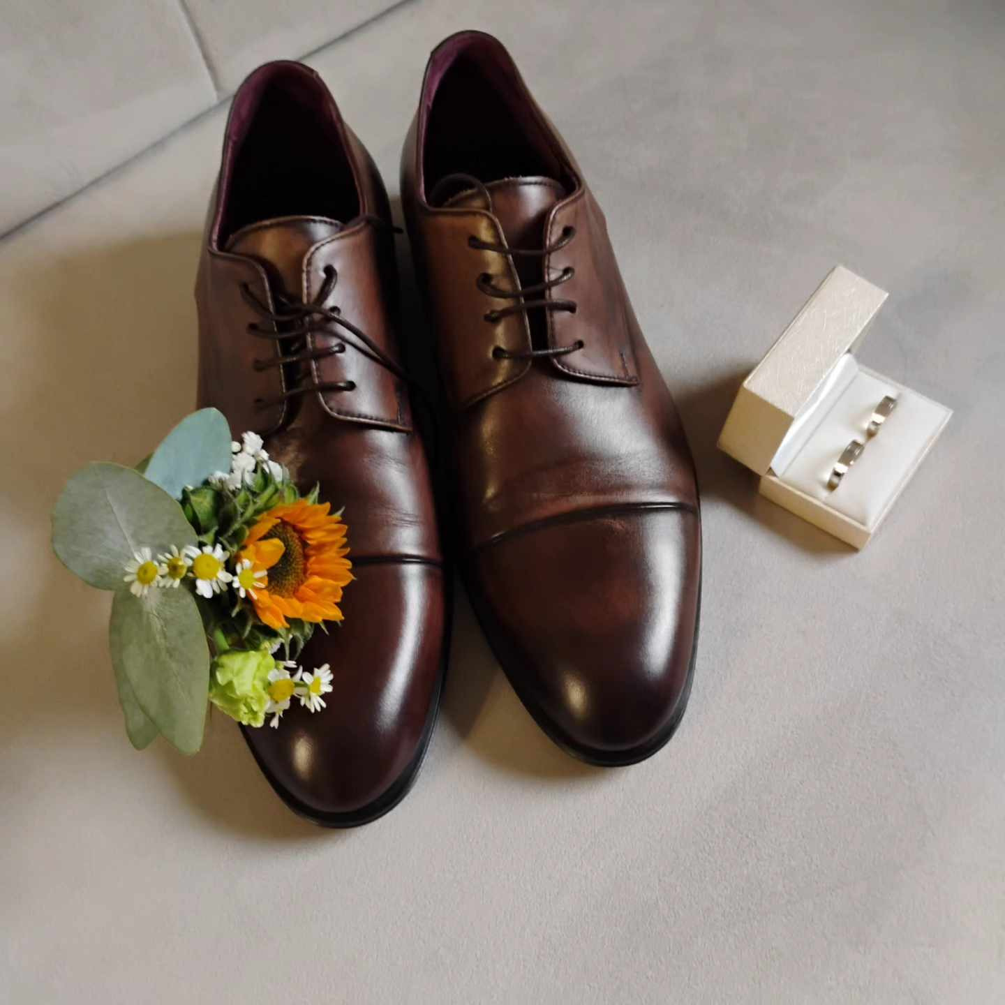 Brązowe skórzane buty męskie z butonierką z małym słonecznikiem i białymi margaretkami, obok pudełko z obrączkami na jasnym tle.