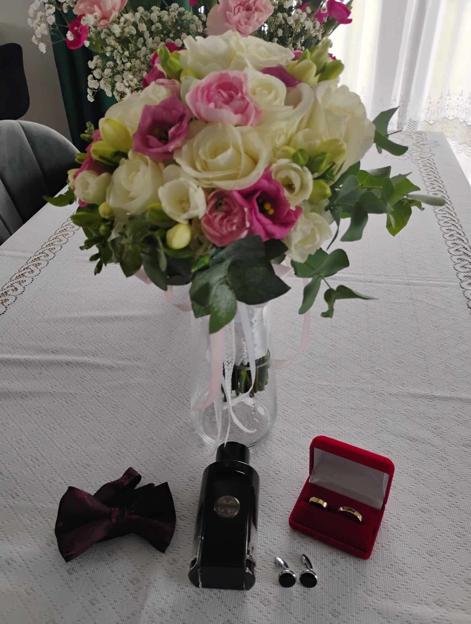 Bukiet ślubny z białych i różowych kwiatów w wazonie, obok bordowa mucha, flakon perfum, spinki do mankietów i czerwone pudełko z obrączkami na białym obrusie z koronką.